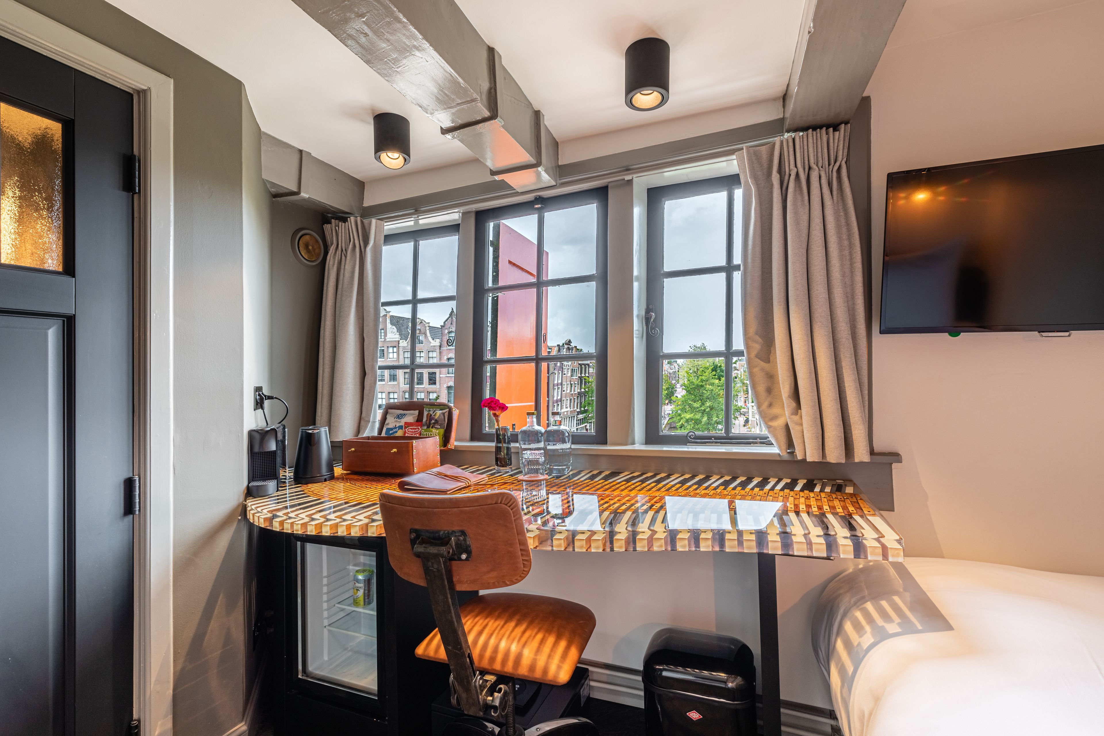 A compact hotel room with a bed, a small desk with a chair, a TV mounted on the wall, and a window with curtains.
