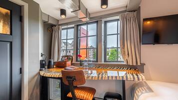 A compact hotel room with a bed, a small desk with a chair, a TV mounted on the wall, and a window with curtains.