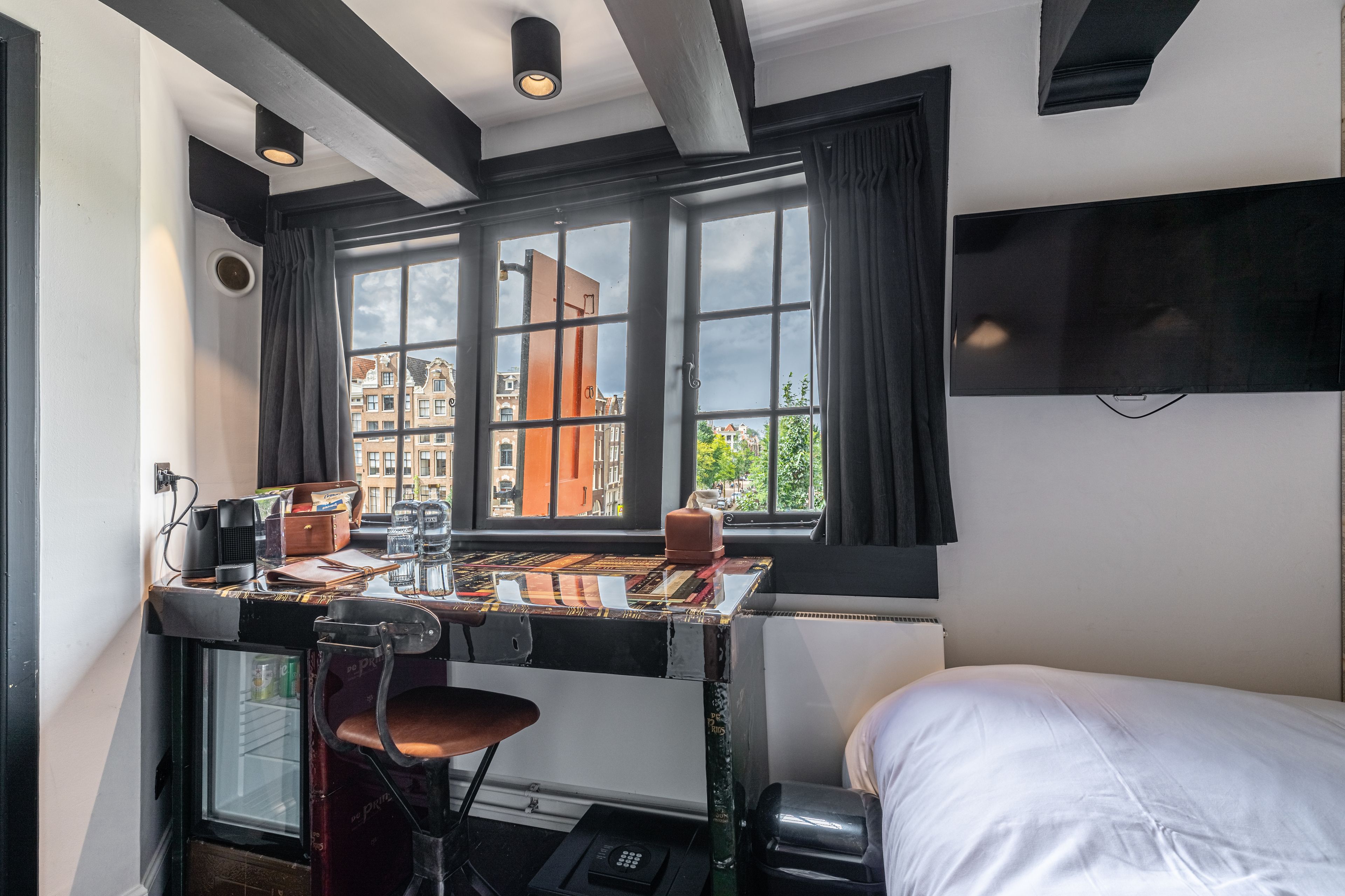 A compact hotel room with a small desk, a chair, a bed, and a large window offering a view of buildings and trees.