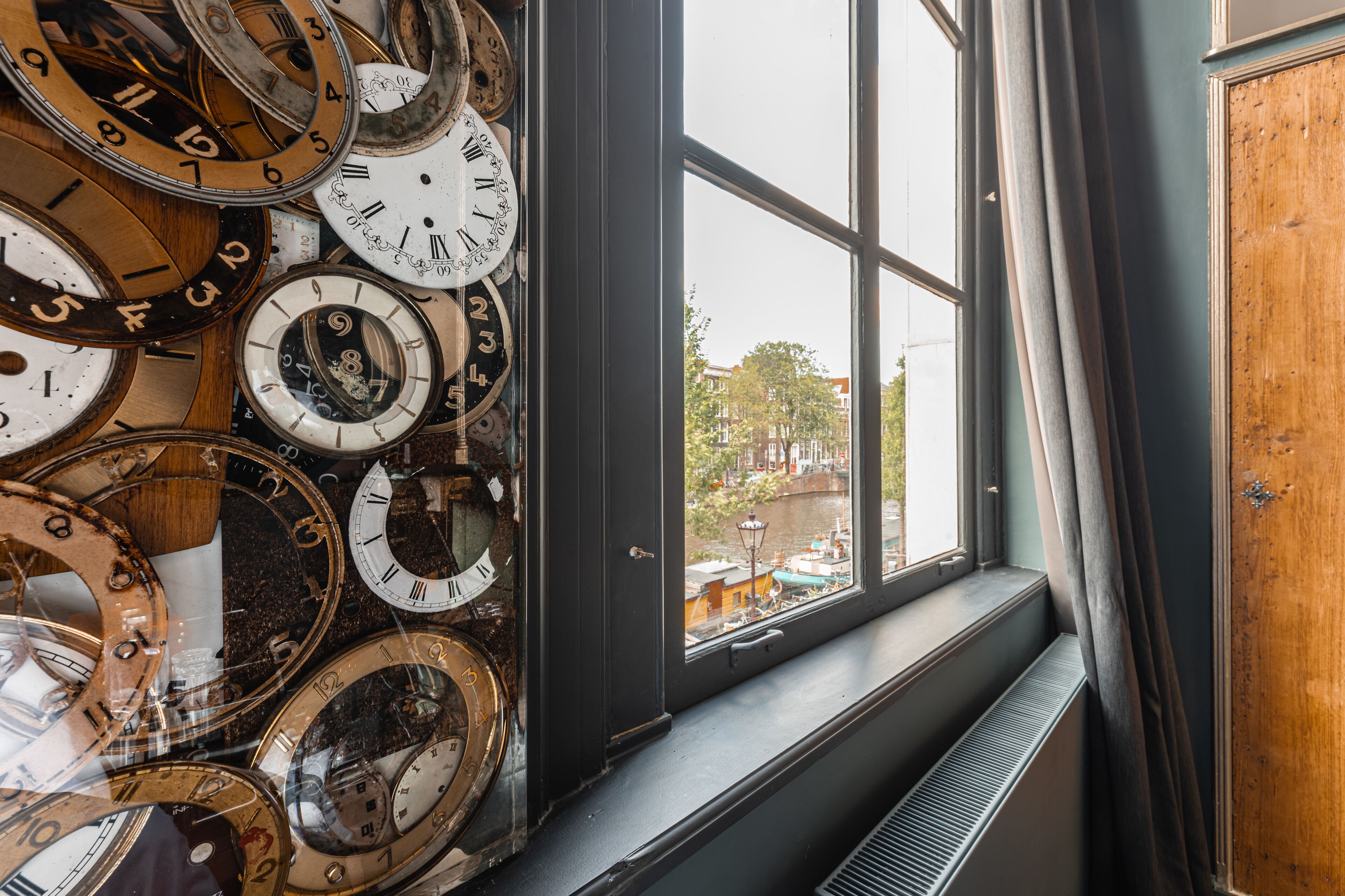 A collection of clocks on a wall, a window with a view of buildings, and a radiator.