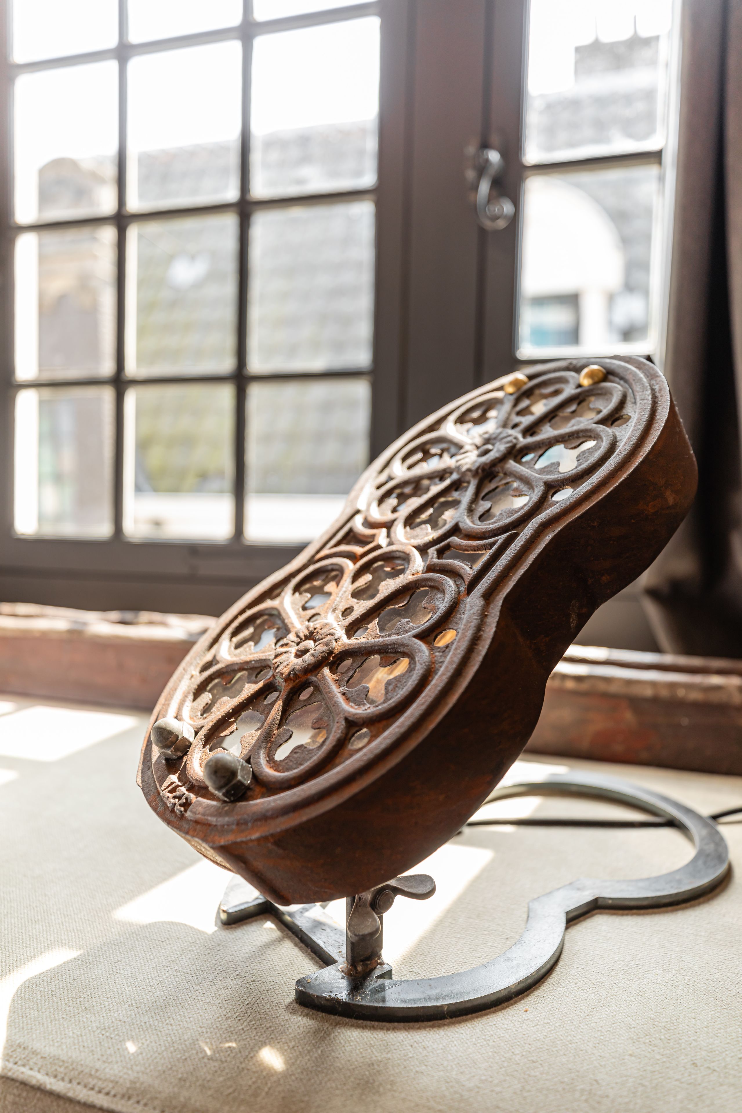 A metal object with intricate designs, possibly a decorative piece, placed on a stand in a room with a large window in the background.