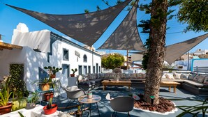 An outdoor seating area with a white building, a tree, and various potted plants. - 1812 Caves 'n Mansion (Santorini)