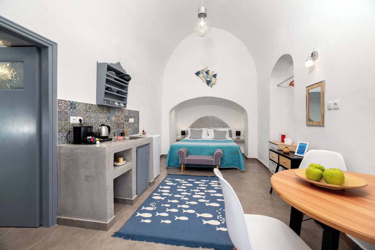 Ein kompakter Raum mit Küchenzeile, einem Bett mit türkisfarbenem Überwurf, einer Essecke mit Holztisch und Stühlen sowie einem blauen Teppich mit Fischmotiven.