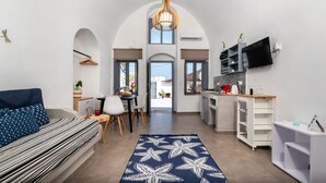 A modern living room with a sofa, dining area, kitchen, and a large arched window. - 1812 Caves 'n Mansion (Santorini)