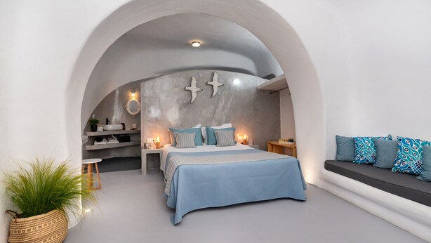 A modern bedroom with a bed, a seating area, and a kitchen in the background. - 1812 Caves 'n Mansion (Santorini)