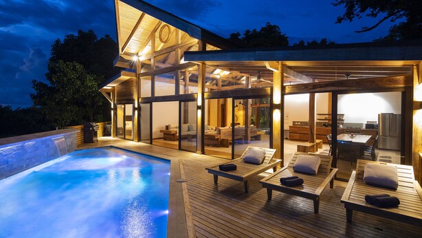 A modern outdoor pool area with wooden deck and lounge chairs, connected to a glass-enclosed living space with a dining area. - Villa Melody (Cóbano)