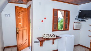 A compact kitchen with a refrigerator, a wooden shelf, a window with shutters, and a television mounted on the wall. - Pousada Vale dos Sonhos (Saquarema)
