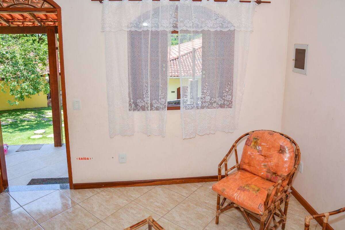 Une pièce avec un fauteuil en osier, une fenêtre ornée de rideaux en dentelle et une vue sur un jardin.