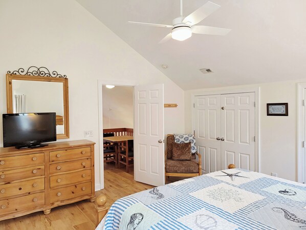 A bedroom with a bed, a dresser, a television, a ceiling fan, and a door leading to a dining area.