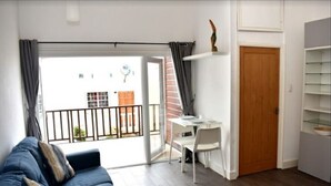 Ein Wohnzimmer mit einem blauen Sofa, einem kleinen weißen Schreibtisch mit Stuhl, einer Holztür und einem Balkon mit Blick auf ein Gebäude.