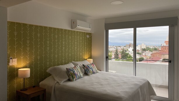 Makuuhuoneessa on sänky, kaksi yöpöytävalaisinta, seinälle asennettu ilmastointilaite ja liukuovesta avautuva näkymä kaupunkimaisemaan.