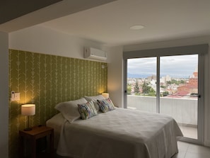 A bedroom with a bed, two bedside lamps, a wall-mounted air conditioner, and a view of a cityscape through a sliding glass door. - Simpatico Loft Centrico (Salta)