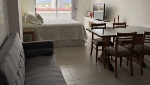 Hotellihuone, jossa on sänky, sohva, ruokapöytä tuoleineen, televisio ja suuret ikkunat, joista avautuu näkymä kaupunkiin.