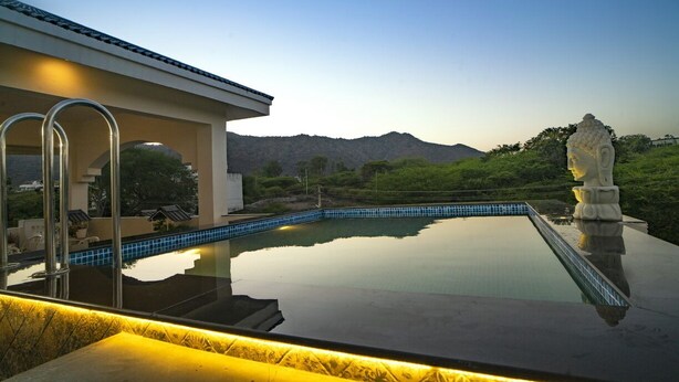 A modern pool with clear blue tiles, a stone sculpture, and a view of mountains in the background. - Anagha Homestay Home Away From Home (Udaipur)