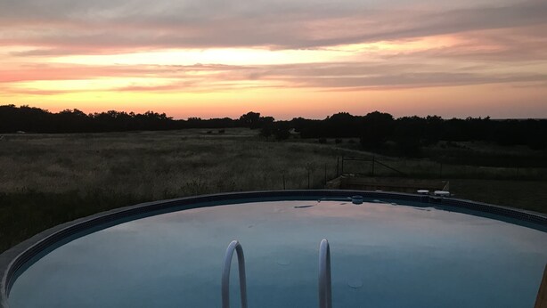 A swimming pool with a wooden deck and metal ladders, surrounded by a grassy field and trees, under a sunset sky. - Your next, beautiful getaway! (Hunt)