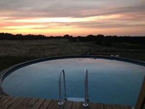 Une piscine dotée d’une terrasse en bois et d’échelles en métal, entourée d’un champ verdoyant et d’arbres, sous un ciel au coucher du soleil.