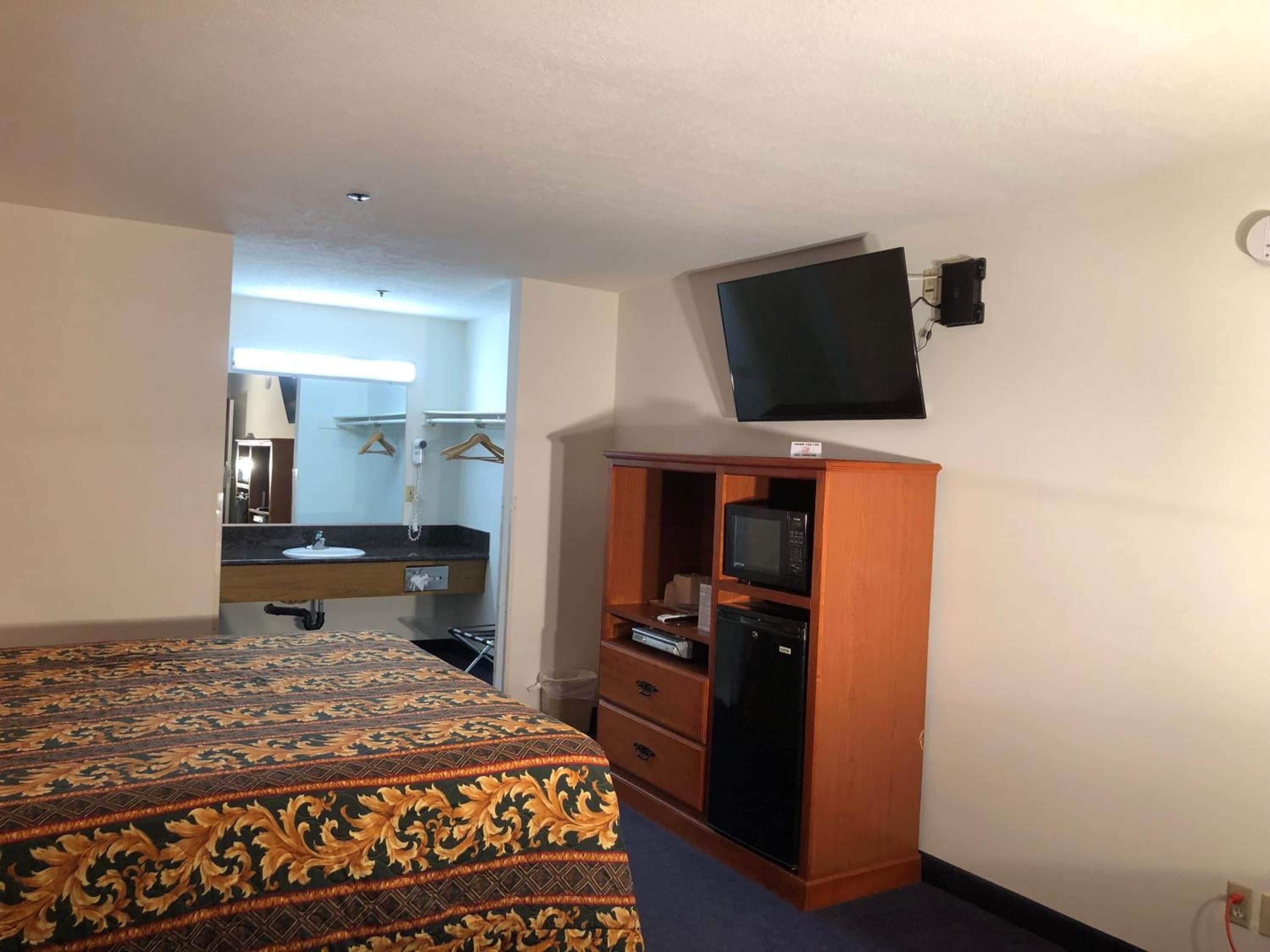 A hotel room with a bed, a wooden wardrobe, a television mounted on the wall, and a bathroom visible through an open door.