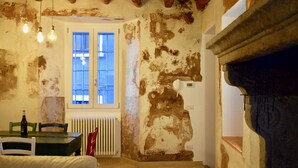 Una habitación rústica con paredes de ladrillo visto, techo de madera, una mesa de comedor con sillas, una ventana con contraventanas y un radiador.