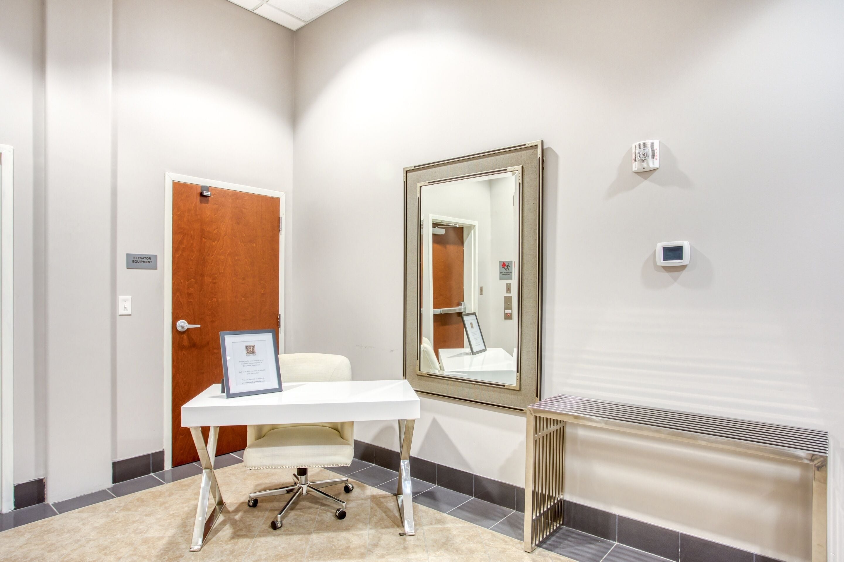 A hotel room with a white desk, a framed certificate, a mirror, a radiator, and a door with a sign.