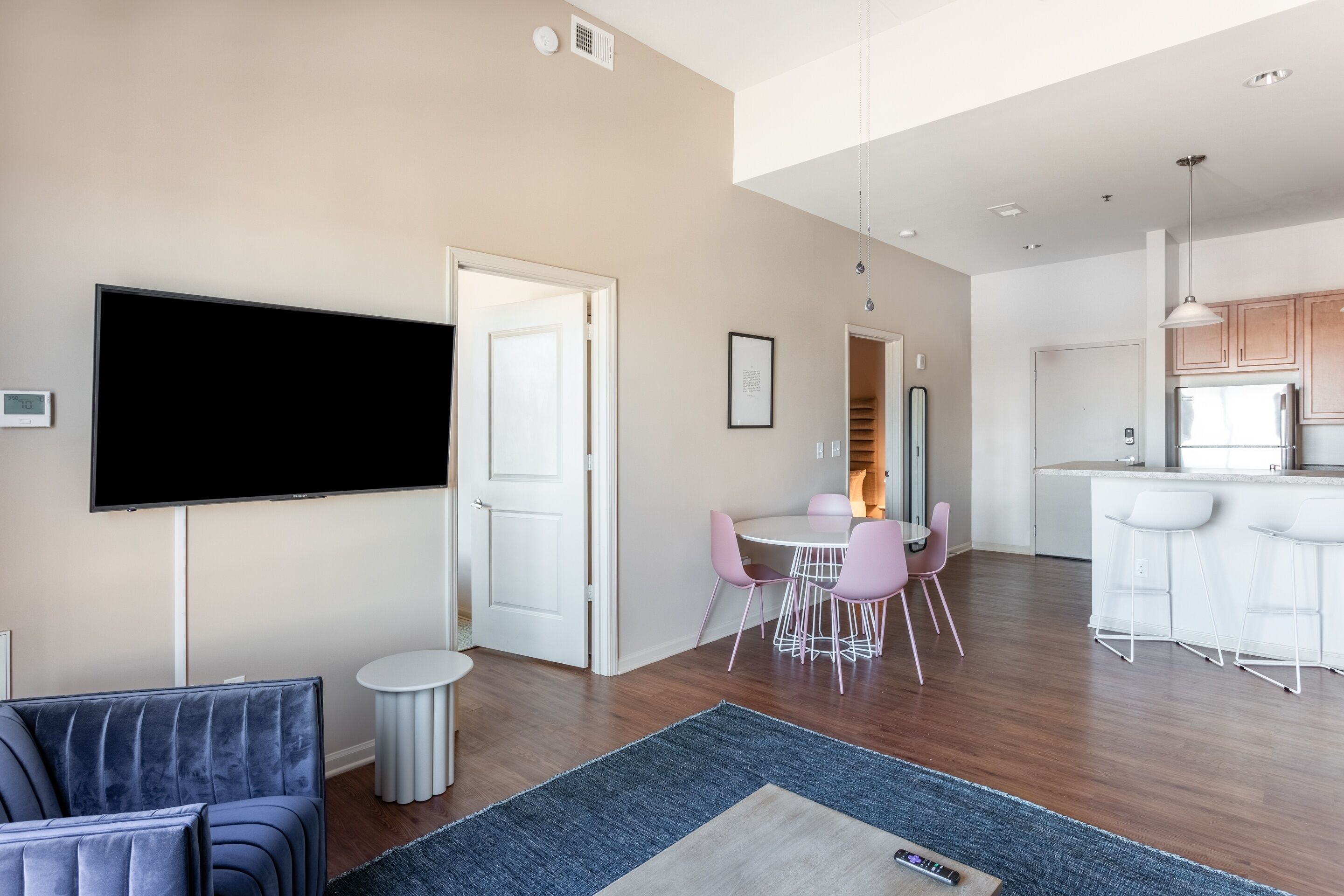 A modern living room with a flat-screen TV, a sofa, a dining area with pink chairs, and a kitchen with white cabinets and a bar.