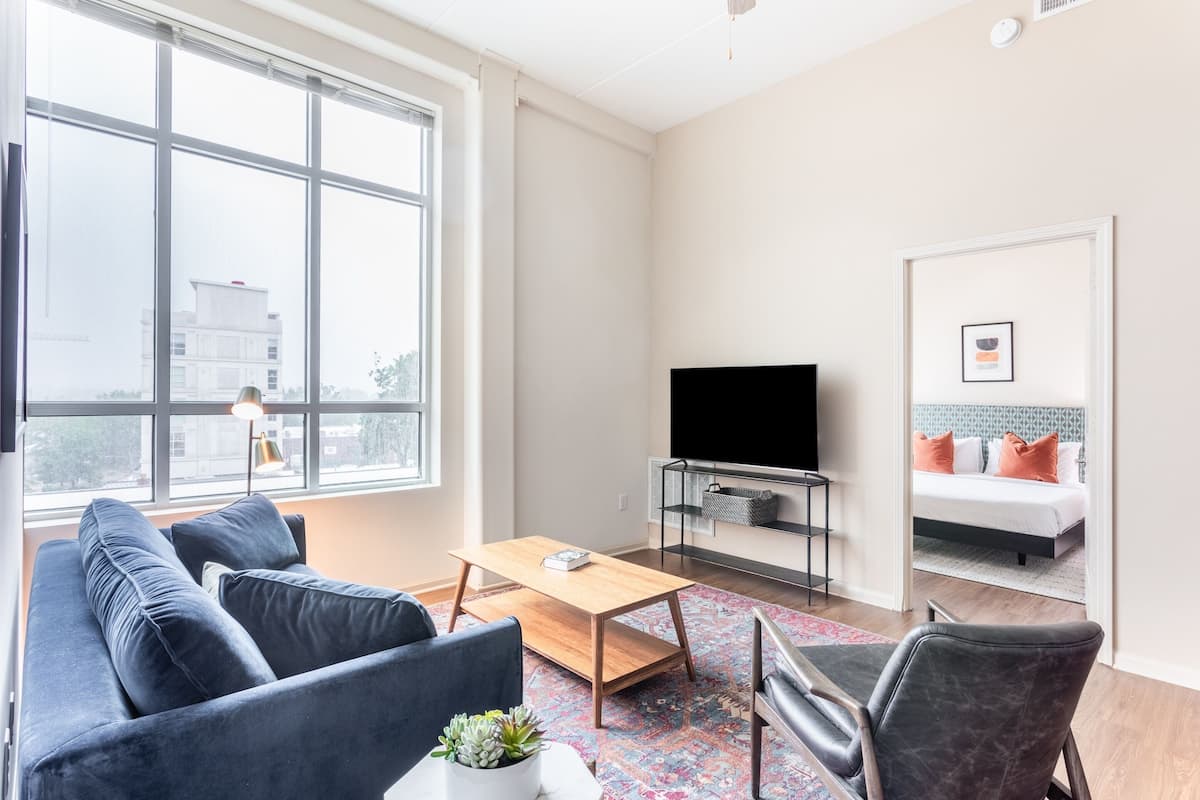 Un salon moderne avec un canapé, une table basse et un téléviseur. On aperçoit un lit avec des coussins orange à travers une porte ouverte.