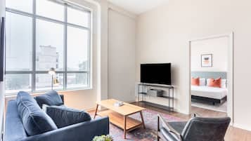 A modern living room with a sofa, coffee table, and TV. A bed with orange pillows is visible through an open door.