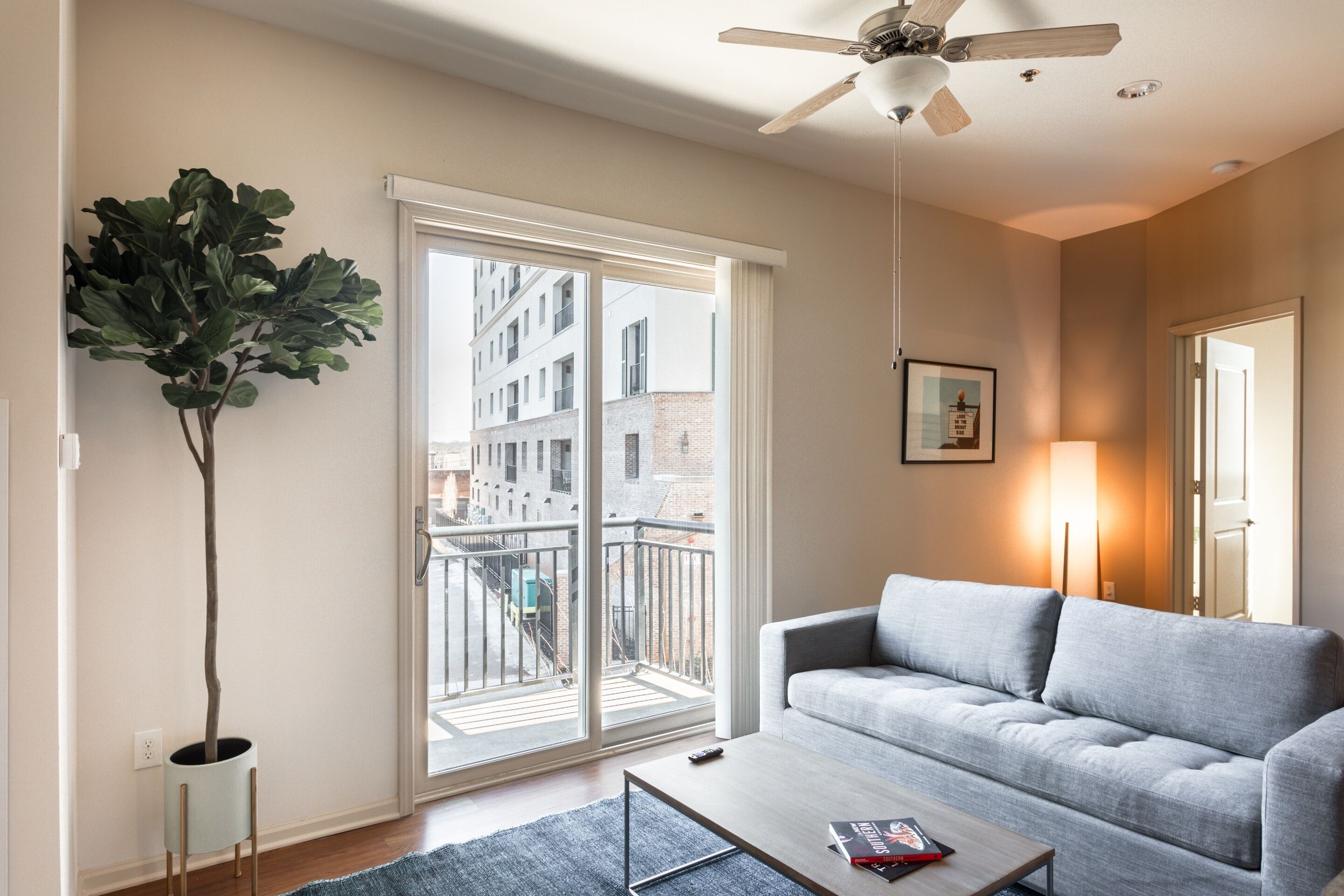 A modern living room with a grey sofa, a coffee table, a large window with a view of buildings, and a ceiling fan.