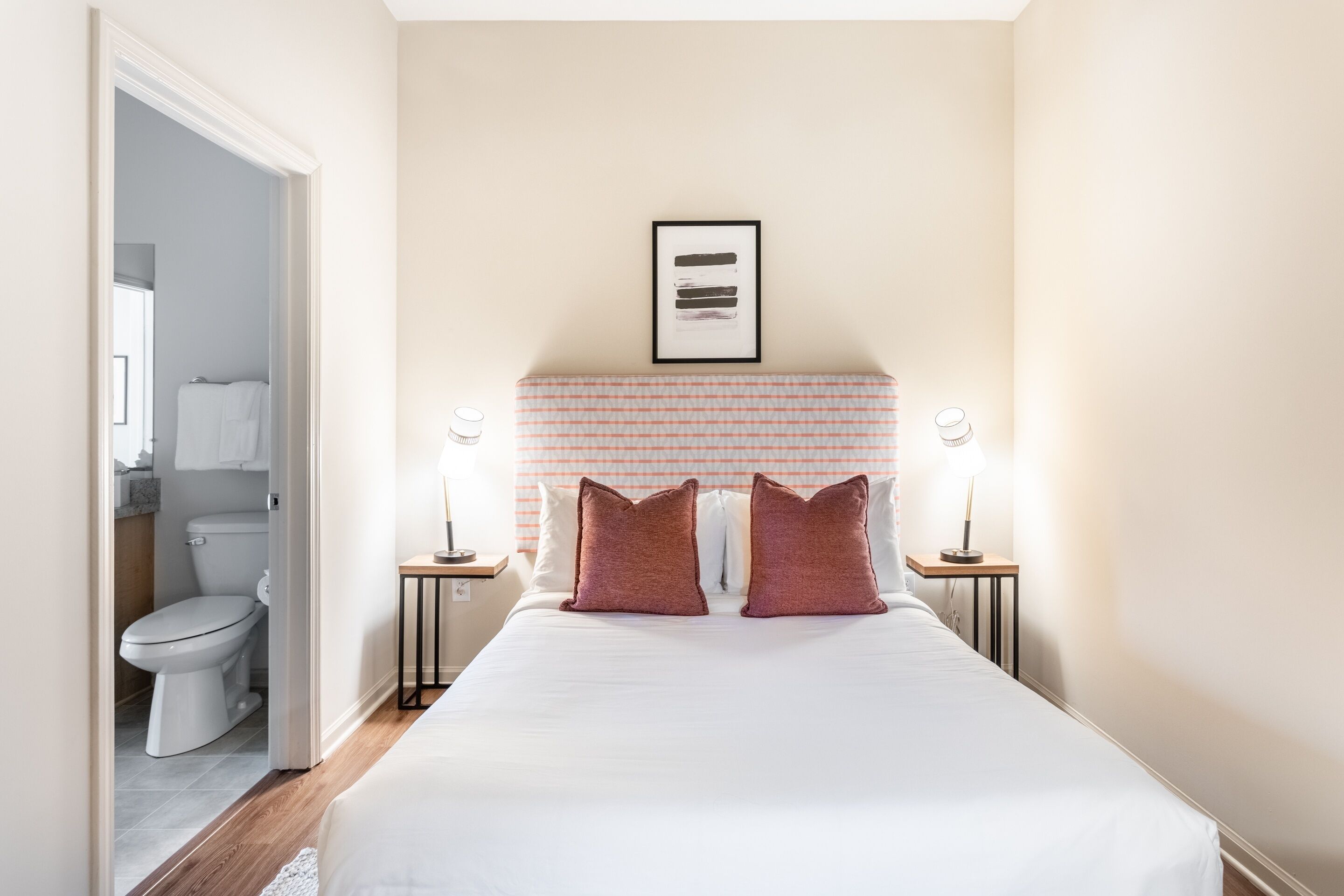 A bedroom with a bed, two bedside lamps, a framed picture on the wall, and a bathroom visible through an open door.