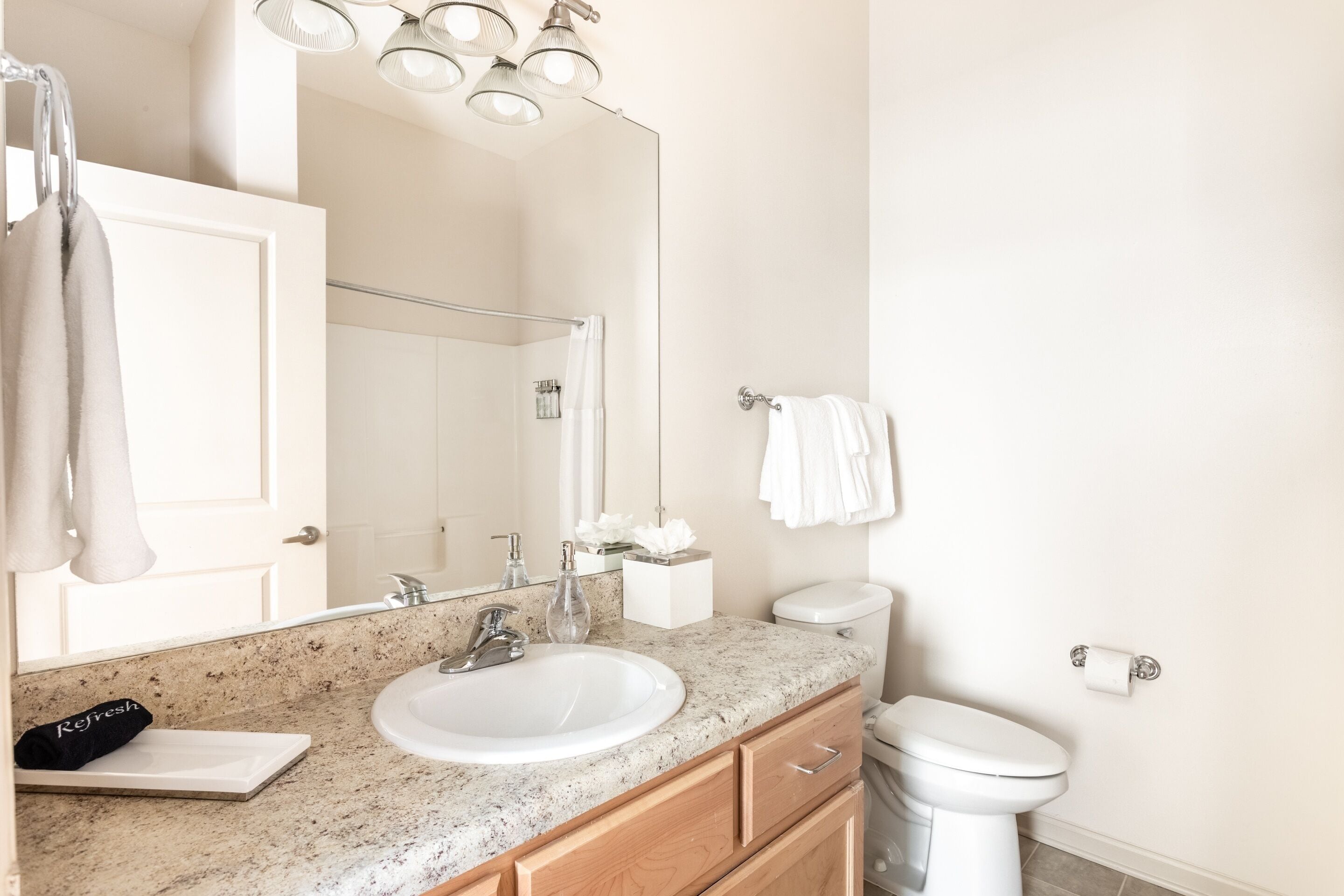A bathroom with a white sink, mirror, and toilet. There are towels hanging on the wall and a trash can next to the toilet.
