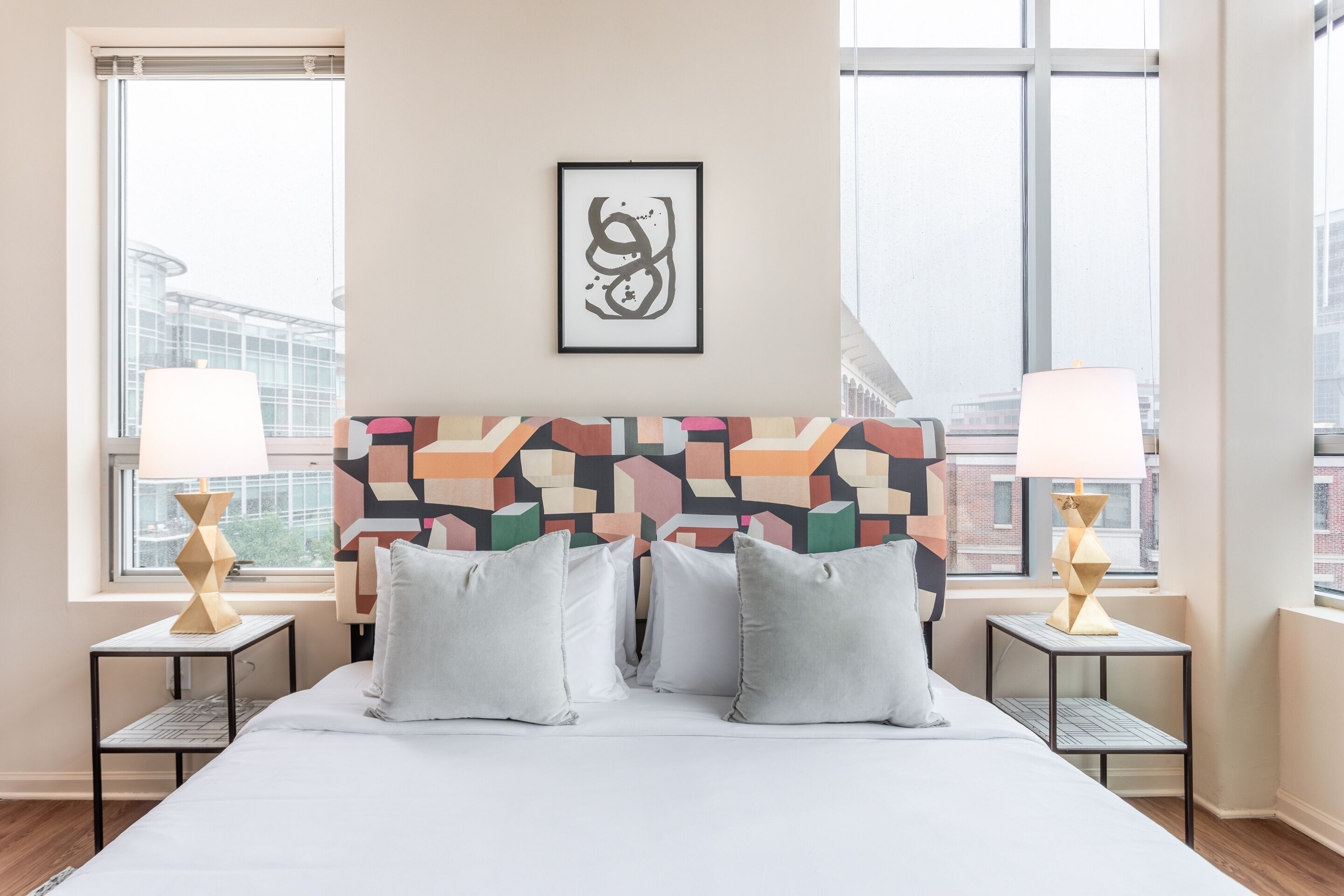 Een moderne hotelkamer met een groot bed, twee nachtkastjes met lampen, een kleurrijk geometrisch hoofdeinde en een ingelijste kunstwerk aan de muur.
