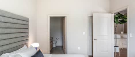 Un dormitorio con una cama, una mesita de noche, una lámpara de noche, una puerta que da a un baño y una planta en una esquina.