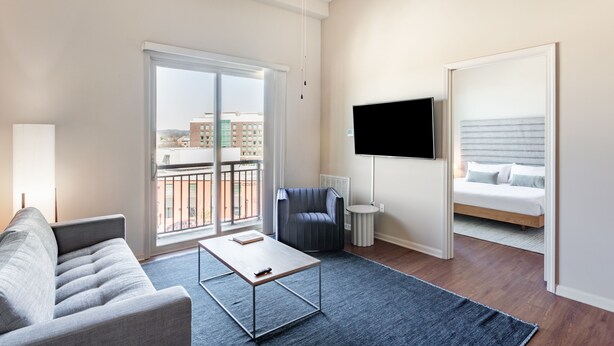 A modern living room with a sofa, a coffee table, a TV, and a balcony with a view of buildings. - Mint House Greenville Downtown by Kasa (Greenville)