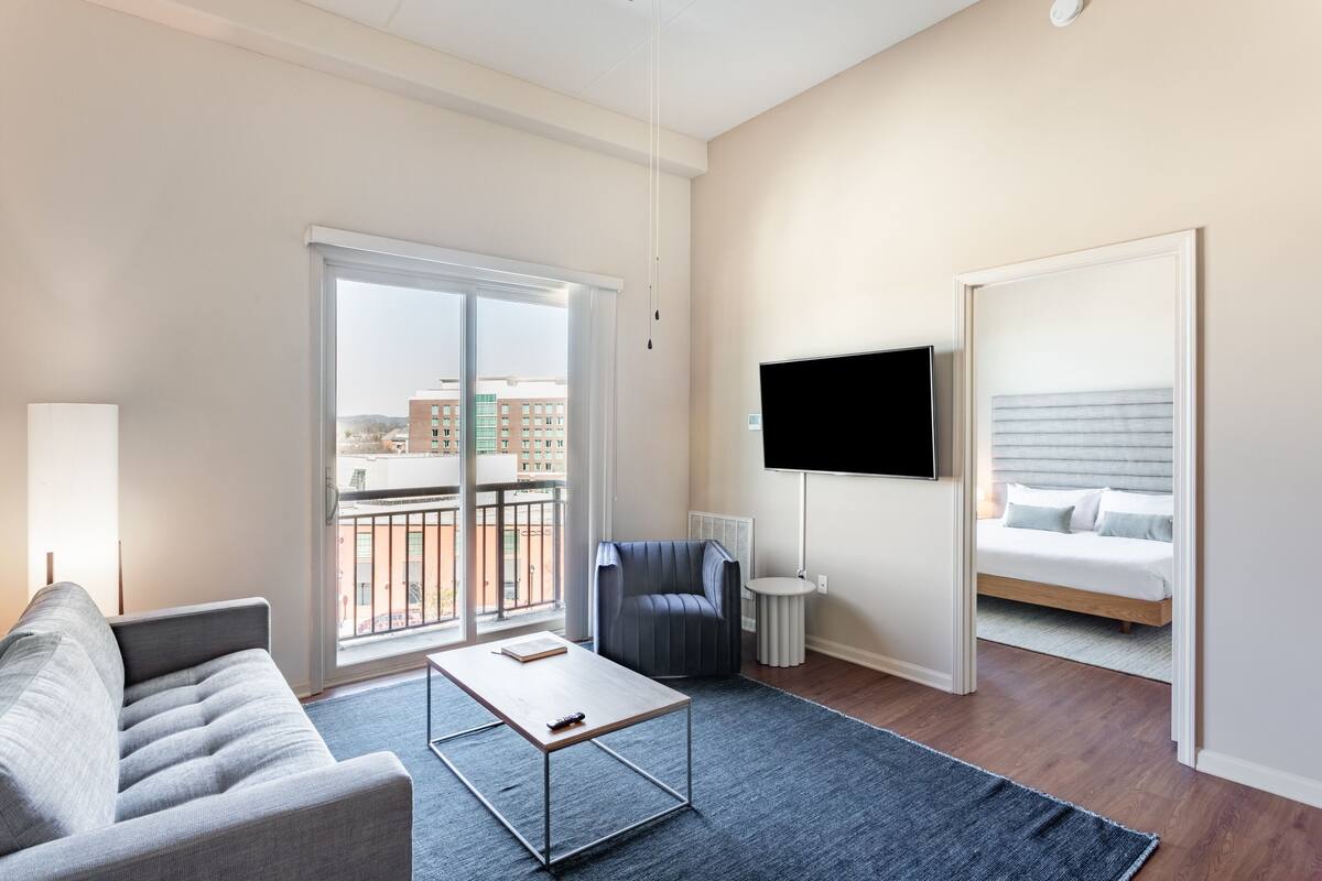 Un salon moderne avec un canapé, une table basse, un téléviseur et un balcon offrant une vue sur les immeubles.