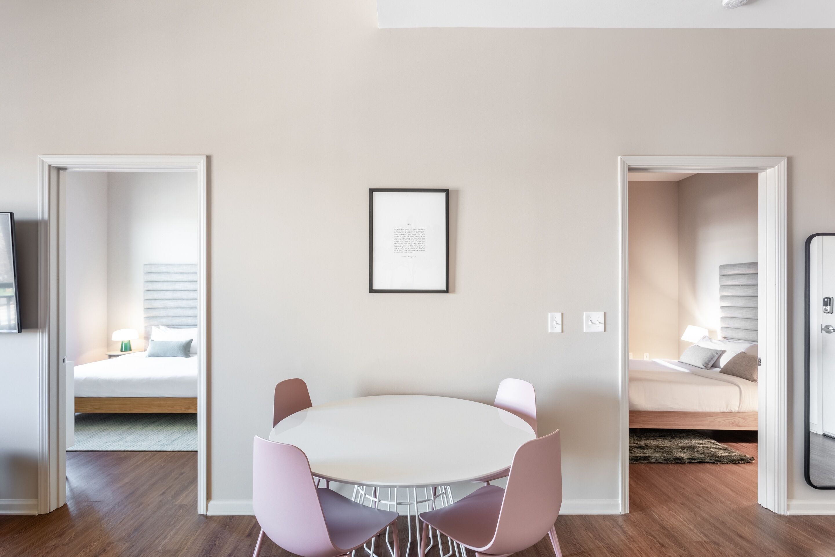 A modern room with a round table and pink chairs, a framed picture on the wall, and a bed in the background.