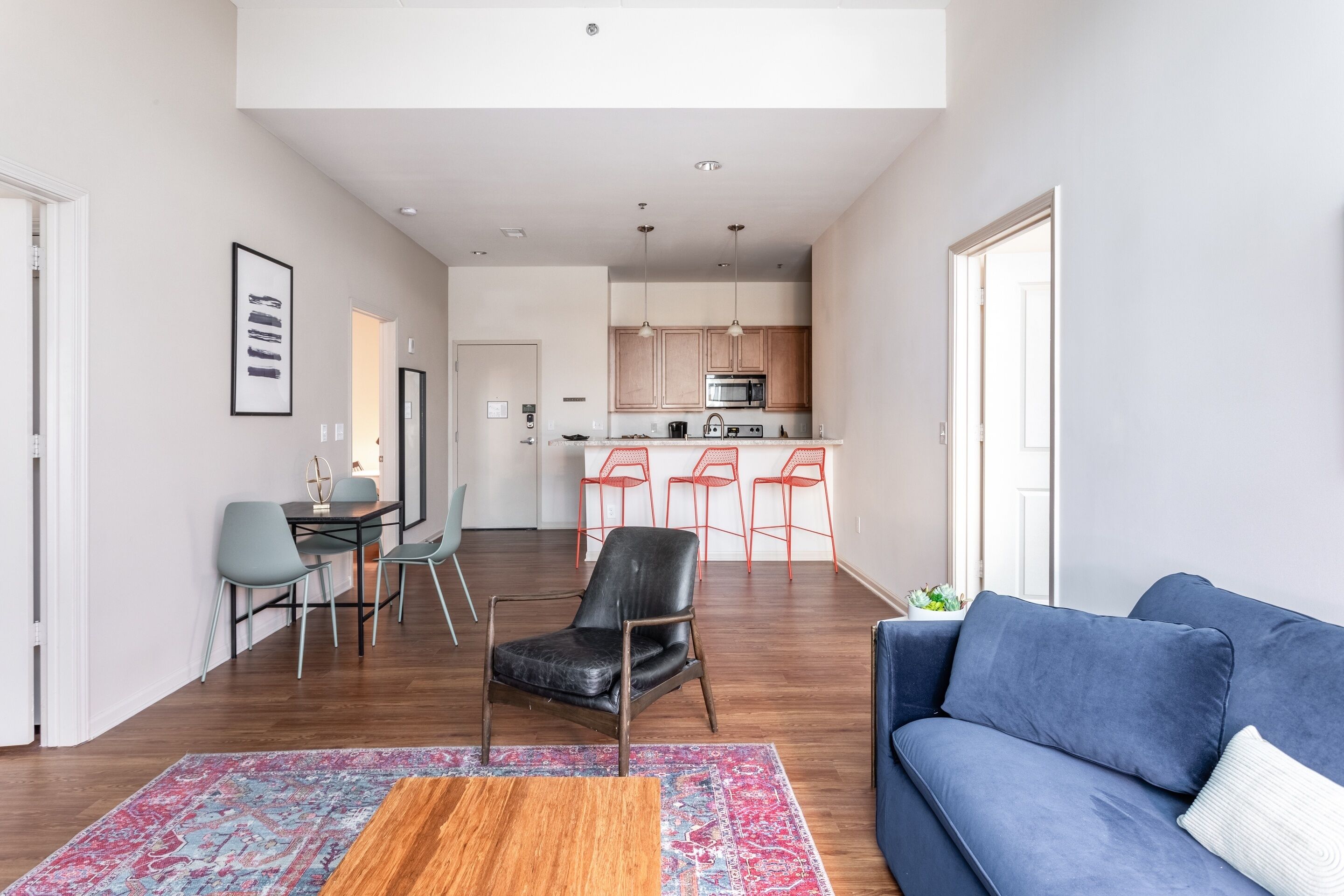 Una sala moderna con un sofá azul, una mesa de centro de madera y un tapete estampado. Se observa una cocina al fondo con un microondas y sillas rojas.