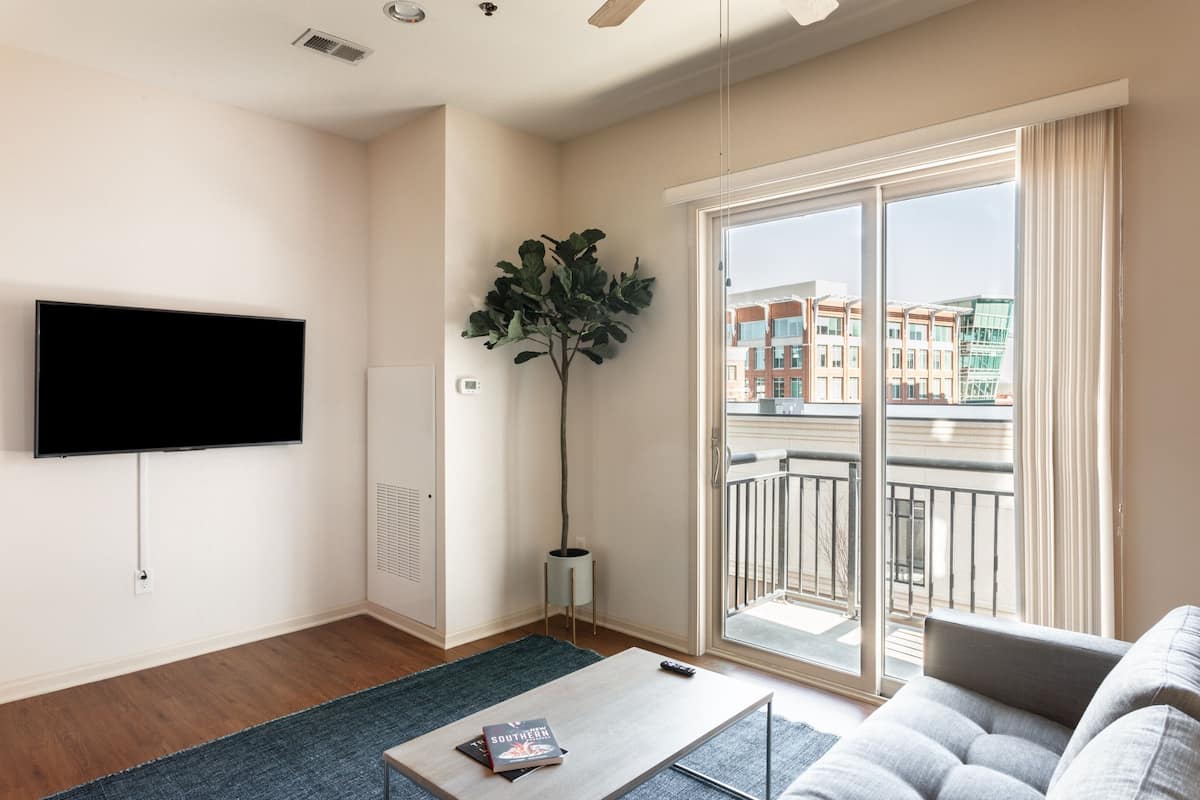Un salon moderne doté d’un téléviseur à écran plat, d’une grande plante, d’un canapé et d’un balcon offrant une vue sur la ville.