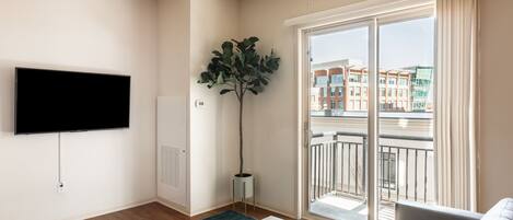 A modern living room with a flat-screen TV, a large plant, a sofa, and a balcony with a city view.