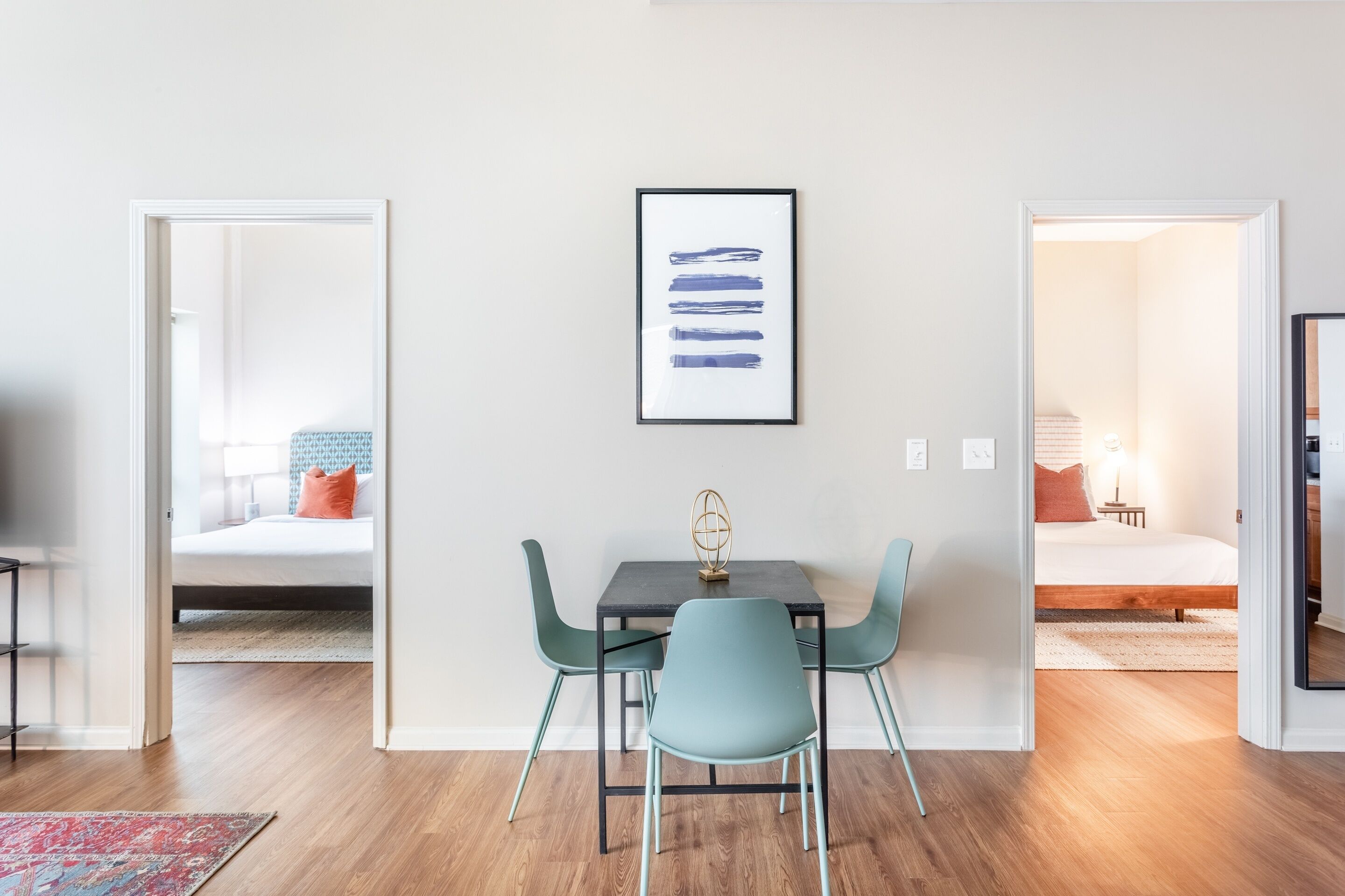 A modern room with a wooden floor, a dark table with teal chairs, a framed artwork, and a bed with a nightstand in the background.