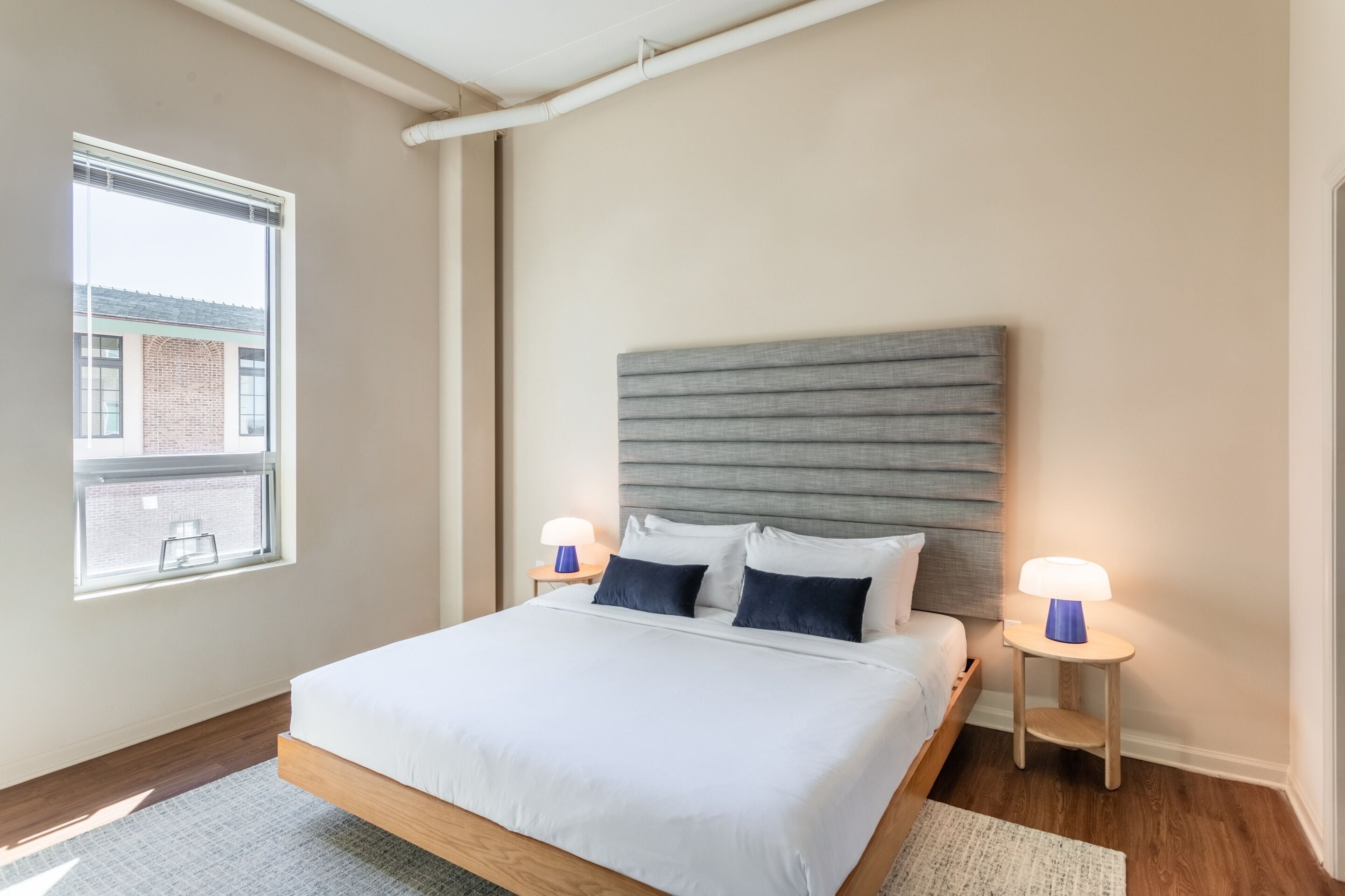 A modern bedroom with a bed, two bedside tables, a window, and a view of a building outside.