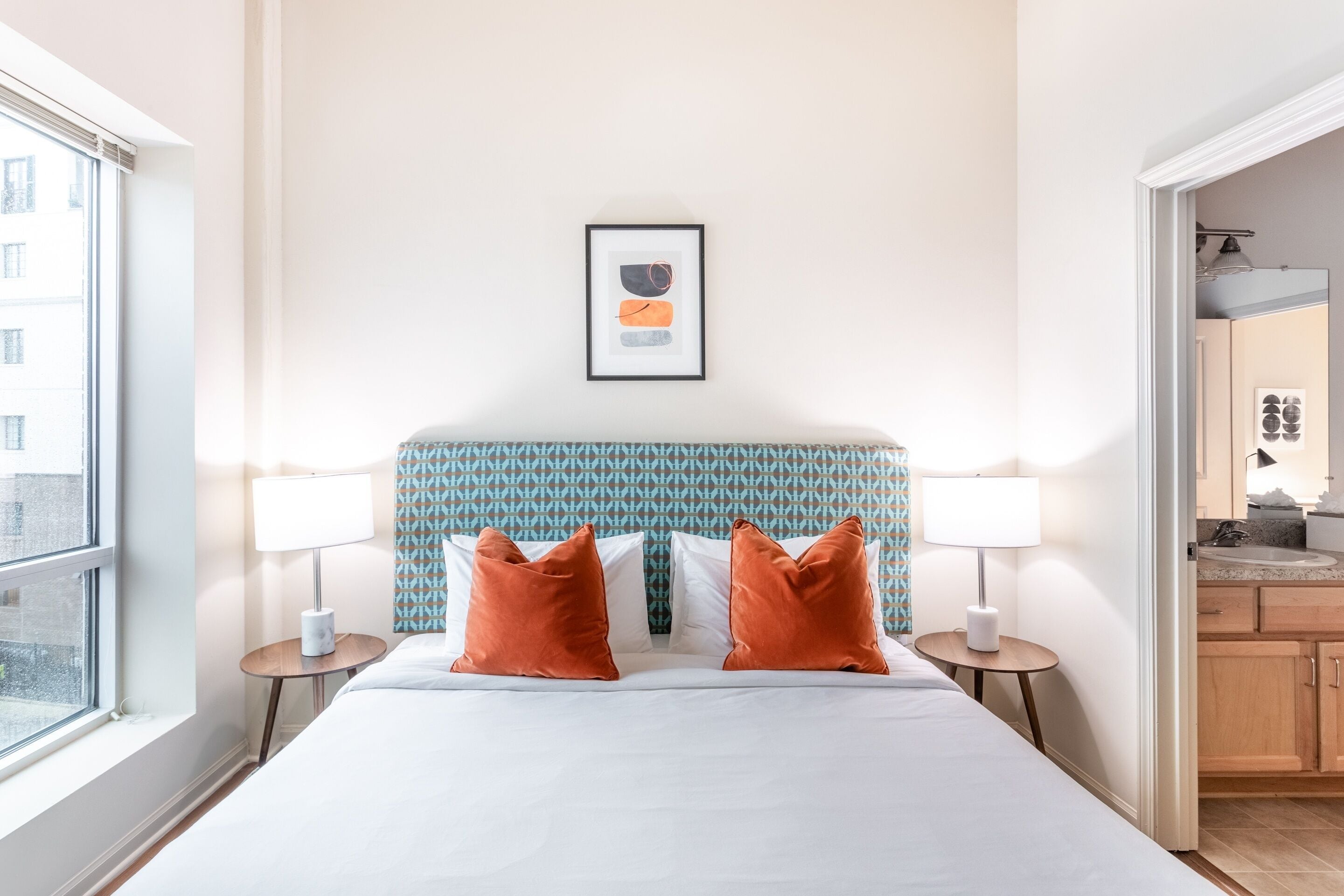 Ein Hotelzimmer mit einem großen Bett, zwei Nachttischen mit Lampen, einem gerahmten Kunstwerk an der Wand und Blick auf die Stadt durch das Fenster.