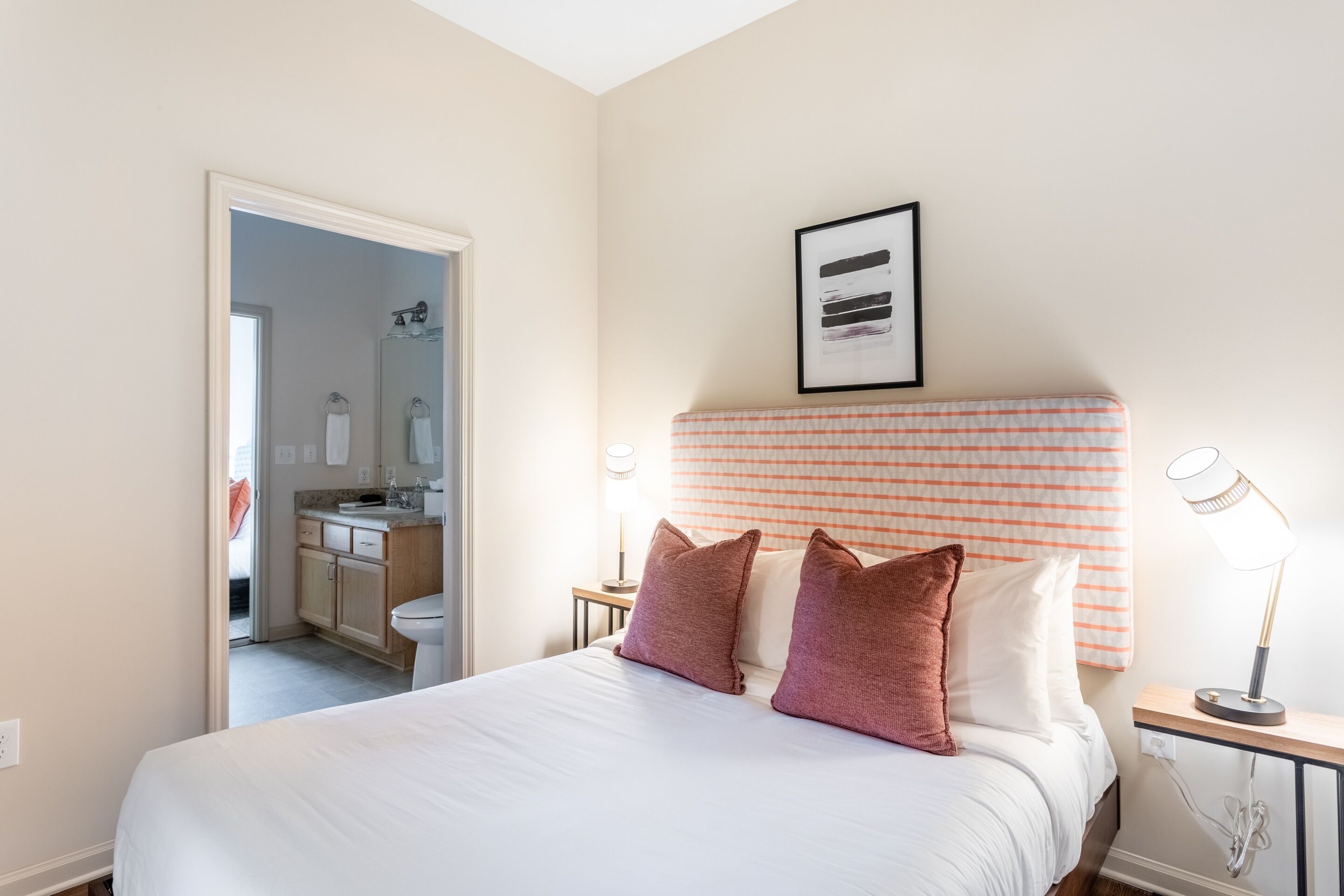 Habitación de hotel con cama, mesita de noche con lámpara, cabecero con estampado a rayas y un cuadro en la pared.