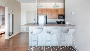 A modern kitchen with wooden cabinets, a stainless steel refrigerator, a microwave, and a granite countertop. There are three white bar stools and two pendant lights. - Mint House Greenville Downtown by Kasa (Greenville)