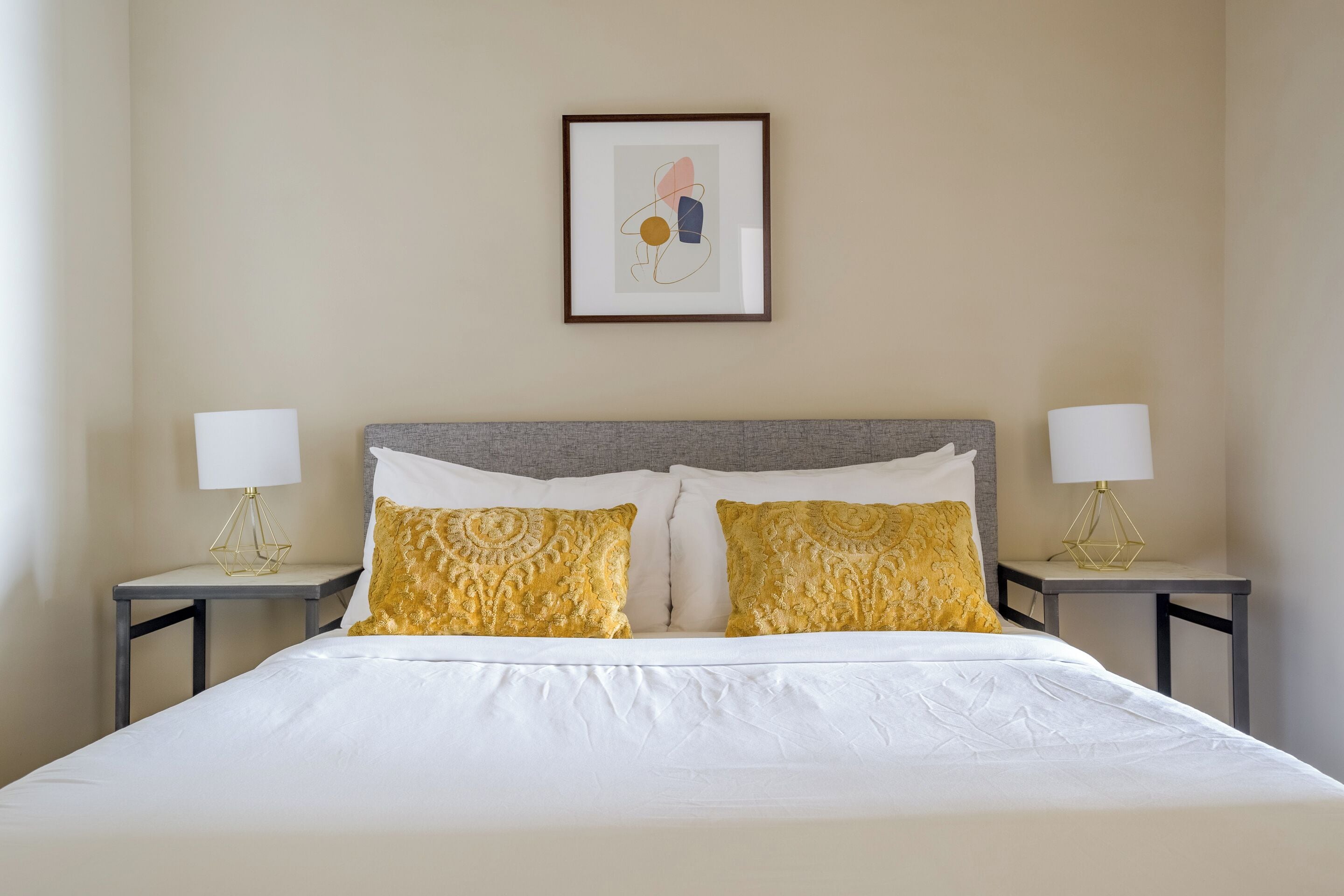 A neatly made bed with white linens and two decorative pillows, flanked by two bedside tables with lamps.
