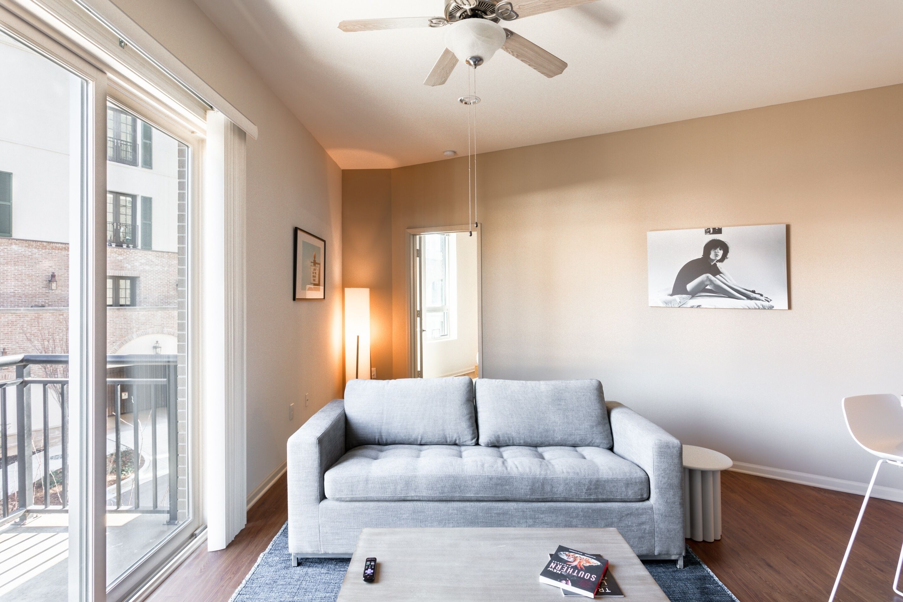 A modern living room with a grey sofa, a coffee table with books, a ceiling fan, and a large window with a view of a building.