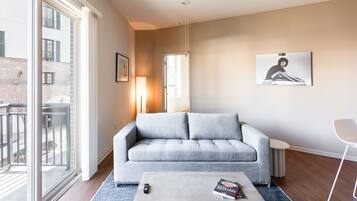 A modern living room with a grey sofa, a coffee table with books, a ceiling fan, and a large window with a view of a building.