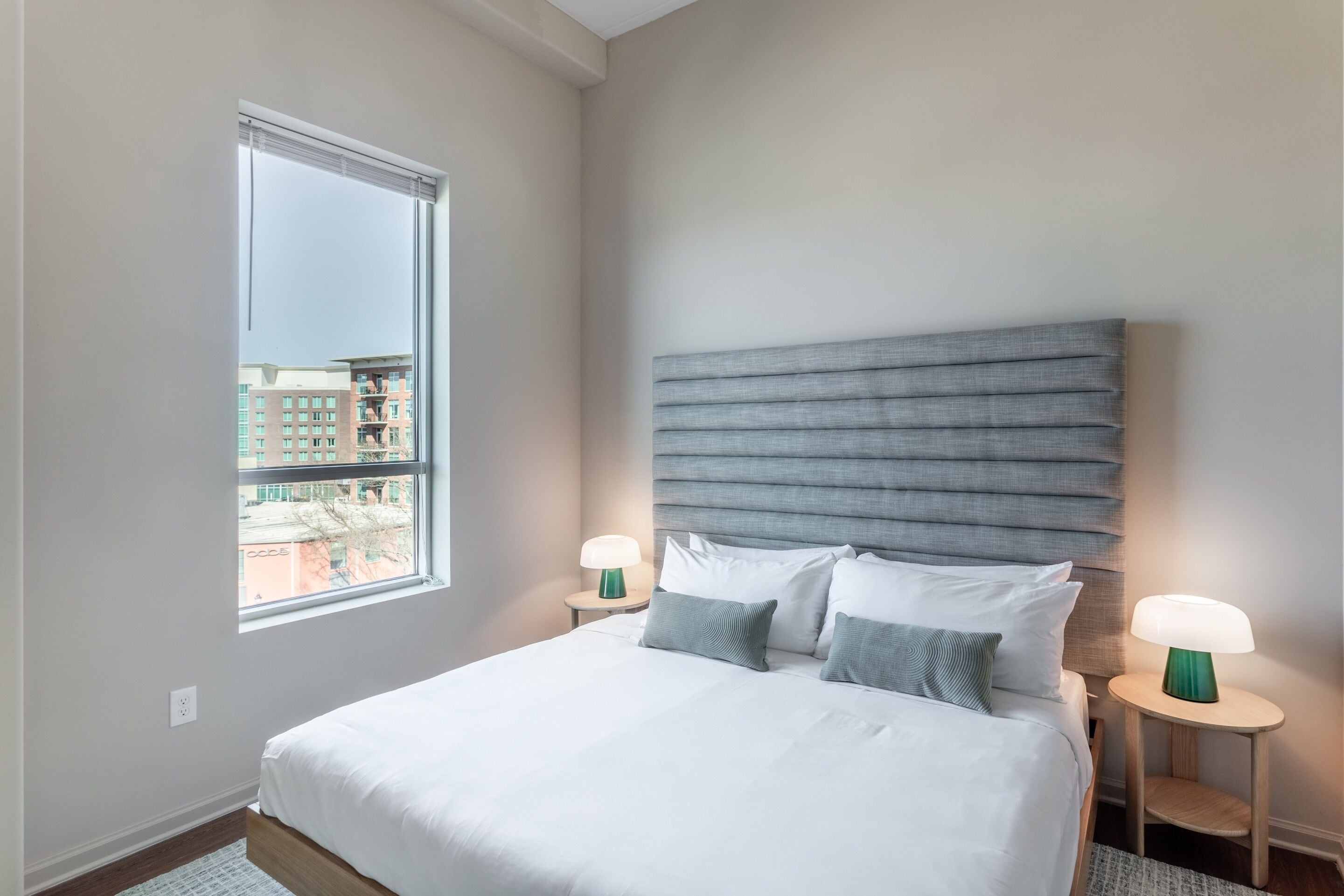 Ein Schlafzimmer mit einem großen Bett, zwei Nachttischen mit Lampen, einem Fenster mit Jalousien und Blick auf Gebäude.