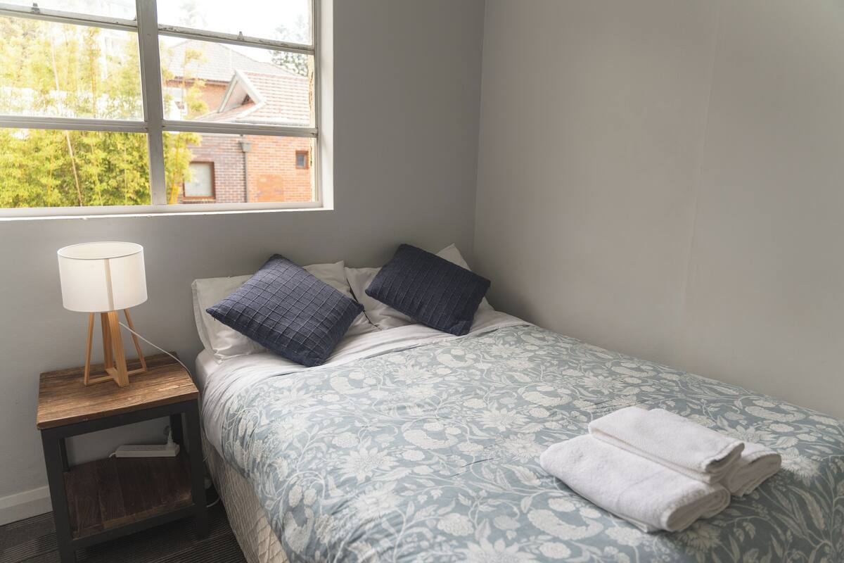 Un dormitorio con cama, almohadas, mesita de noche, lámpara y ventana con vista a edificios y árboles.