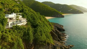 A modern house with multiple levels, situated on a lush green hillside overlooking a clear blue sea. - 5 bedroom villa with pool in stunning clifftop location next to gorgeous beach (Rogues Bay)