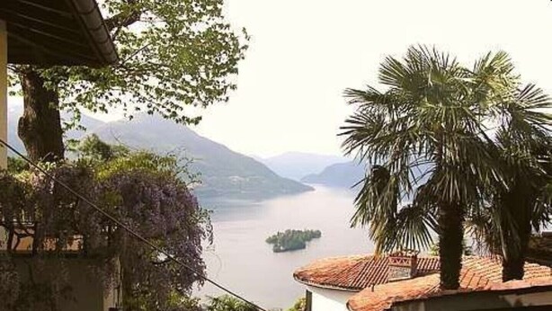Blick auf einen See mit Bergen im Hintergrund, eine Palme und ein Gebäude mit einem Ziegeldach.
