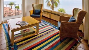 Ein Zimmer mit großem Fenster, Blick auf Palmen, ein Korbstuhl, ein Holztisch und ein farbenfroher Streifen Teppich.